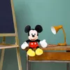 A Mickey Mouse plush toy sits on a wooden desk next to a yellow desk lamp and some pencils, with a chalkboard easel in the background. Voir les images du produit