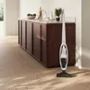 A white cordless vacuum cleaner stands on a wooden floor in front of a dark wood cabinet. Voir les images du produit