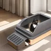 A black and white cat is sitting in a modern, open-top litter box with a ramp. The litter box is placed on a floor with sunlight streaming in from a window. Produktbild-Vorschau