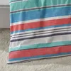 A close-up view of a striped fabric, likely a blanket or bedding, with a textured weave. The fabric is laid out on a soft, fuzzy surface, possibly a rug or carpet, and partially rests against a light-colored, upholstered piece of furniture. Produktbild-Vorschau