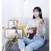 A young woman is sitting on a couch and playing a ukulele, while a selfie stick with two phones is positioned in front of her, showing her from different angles. Podgląd zdjęcia produktu