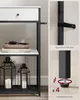A close-up view of a narrow console table with a marble-patterned top and black metal frame, showing details of a drawer, shelves with books and lanterns, and safety features like an anti-tip strap and adjustable feet. Produktbild-Vorschau