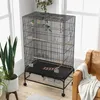 A black metal bird cage with wooden perches and toys, placed indoors near a window, with a leather armchair and plants. Náhled obrázku produktu