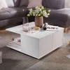 A white coffee table with storage is placed in a living room setting, next to a gray sectional sofa. The table has a vase of white flowers, a pitcher of pink liquid, a glass, and a cookie on top. Magazines are visible in an open shelf on the side of the ta Voir les images du produit