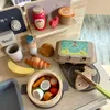 A collection of wooden toy food items, including a croissant, banana, bread roll, egg carton, and a pot with vegetables, arranged on a wooden surface. Produktbild-Vorschau