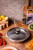 A frying pan with a glass lid is placed on a wooden table, surrounded by ingredients like lemons, tomatoes, and carrots, along with a cookbook and a bottle of oil. Náhled obrázku produktu