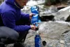 A person is filtering water from a stream into a bottle. Produktbild-Vorschau