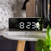 A digital alarm clock with a white LED display sits on a marble-patterned surface next to a blurred background. Podgląd zdjęcia produktu
