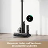 A cordless vacuum cleaner with its charging dock and accessories, displayed against a light-colored wall and floor. Produktbild-Vorschau