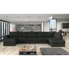 A large, dark gray sectional sofa is shown in a modern living room setting with a bookshelf and large windows in the background. Náhľad obrázku produktu
