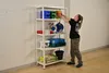 A woman is organizing items on a white metal shelving unit in what appears to be a workshop or storage area. Produktbild-Vorschau