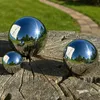 Three polished stainless steel garden spheres of different sizes are placed on a weathered wooden stump. A paved garden path and green grass are visible in the background. Produktbild-Vorschau