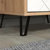 A close-up view of the bottom section of a cabinet, showing its wooden side, a white drawer front with a decorative pattern, and black metal legs resting on a wooden floor. Voir les images du produit