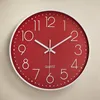 A red wall clock with white numbers and hands is mounted on a textured light beige wall. Náhled obrázku produktu