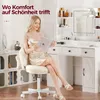 A woman is sitting on a fluffy white chair in front of a white vanity table, reading a book. Náhled obrázku produktu
