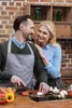 A couple is preparing food in a kitchen. The man is wearing an apron and chopping vegetables on a cutting board, while the woman stands behind him, resting her hand on his shoulder and smiling. Produktbild-Vorschau