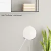 A white router holder is mounted on a light gray wall, with a power cable connected. A plant is visible on the right side of the image. A shelf with books is visible at the top. Náhled obrázku produktu