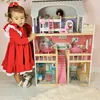 A young girl in a red dress is standing next to a large wooden dollhouse. Voir les images du produit