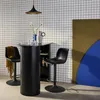 A modern bar area with a high table and two stools, set against a grid-patterned wall and colorful geometric wall art. Produktbild-Vorschau