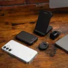 A collection of electronic accessories, including a phone, power bank, wireless charger, and wall adapter, are arranged on a wooden surface. Produktbild-Vorschau