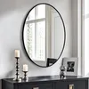 A round mirror with a black frame hangs on a white wall above a dark dresser. Two silver candle holders with white candles are on the dresser to the left of the mirror. A framed picture and a small decorative sculpture are on the dresser to the right of th Podgląd zdjęcia produktu