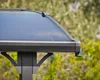 Close-up of the corner of a dark gray carport roof with a translucent dark blue panel, against a blurred green and brown background of foliage. Voir les images du produit