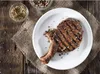 A grilled steak seasoned with peppercorns on a white plate, placed on a burlap cloth on a wooden table. Two small bowls of spices are visible in the background. Podgląd zdjęcia produktu