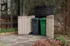 Two trash bins are stored in a Keter garden box on a stone patio, next to a building with large windows and some greenery. Voir les images du produit