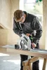 A man is using a circular saw on a wooden plank, which is supported by sawhorses. The scene appears to be a workshop or construction site. Podgląd zdjęcia produktu
