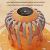 An orange and gray octopus-like head massager on a light orange surface with some small white clips scattered around. Produktbild-Vorschau