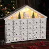 A white wooden advent calendar shaped like a house, decorated with miniature Christmas trees and houses, illuminated by fairy lights, set against a festive background with a Christmas tree and red berries. Voir les images du produit