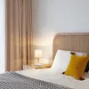 A bedside table with a lamp and pillows next to a bed. Náhľad obrázku produktu