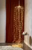 A decorative LED light curtain hangs from the ceiling, cascading down in a waterfall effect. It is placed in a room with a dark red curtain in the background and a small side table with books on the right. Produktbild-Vorschau