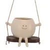 A cute, round, beige planter with a smiling face, sitting on a wooden swing, suspended by rope. Anteprima dell’immagine prodotto