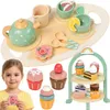 A young girl is holding a wooden flower cookie while a wooden tea set and tiered cake stand are displayed. Produktbild-Vorschau