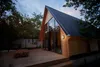 A modern A-frame cabin with large glass doors and windows, set in a wooded area with a wooden deck and outdoor furniture. Voir les images du produit