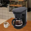 A black coffee maker sits on a wooden table next to a cup of coffee, with a blurred cafe background. Produktbild-Vorschau