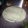 A round rug with a distressed green and brown pattern is placed on a dark wooden floor in a living room setting. A green armchair and a decorative gold tray with a cup are visible in the background. Produktbild-Vorschau