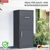 A dark gray parcel box with a mailbox slot and a door, placed against a white wall next to lush green plants and a window. Náhled obrázku produktu