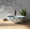 A decorative basin on a wooden countertop with a faucet and a plant in the background. Náhled obrázku produktu