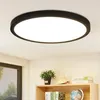 A modern, round, flat LED ceiling light fixture is shown mounted on a white ceiling. Below the light, a wooden shelf holds decorative items including a potted plant, a shell, and books.