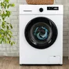 A white washing machine with a black door, showing clothes and water inside, placed in front of a white brick wall and a plant. Produktbild-Vorschau