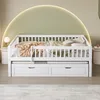 A white wooden daybed with a trundle and drawers, decorated with blankets and pillows, set against a wall with a curved green and yellow light. Produktbild-Vorschau