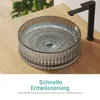 A close-up view of a modern bathroom sink with water swirling down the drain, placed on a wooden countertop. A black faucet is visible to the right. Produktbild-Vorschau