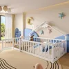 A child's bedroom with a white wooden bed frame designed like a house, complete with a railing and a small fence. Toys and bedding are visible on the bed. The room is decorated with stars and a crescent moon on the wall, and a large window overlooks a gree Podgląd zdjęcia produktu