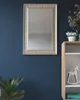 A rectangular mirror with a gold frame hangs on a dark blue wall next to a wooden shelf with decorative items. Náhled obrázku produktu