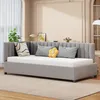 A grey upholstered daybed with a white mattress and several decorative pillows is shown in a well-lit room with a large window and sheer curtains. Produktbild-Vorschau