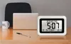 A digital clock with calendar display sits on a wooden desk next to a laptop and a mug. Voir les images du produit