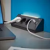 A black power strip with multiple outlets and USB ports is plugged into a laptop on a desk, with a blue wall in the background. Produktbild-Vorschau