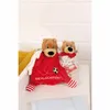 Two teddy bear plush toys, one larger and one smaller, are placed in a basket. The larger teddy bear is attached to a red security blanket with white stripes on its feet. The smaller teddy bear is wearing a red collar and holding a rattle. Produktbild-Vorschau